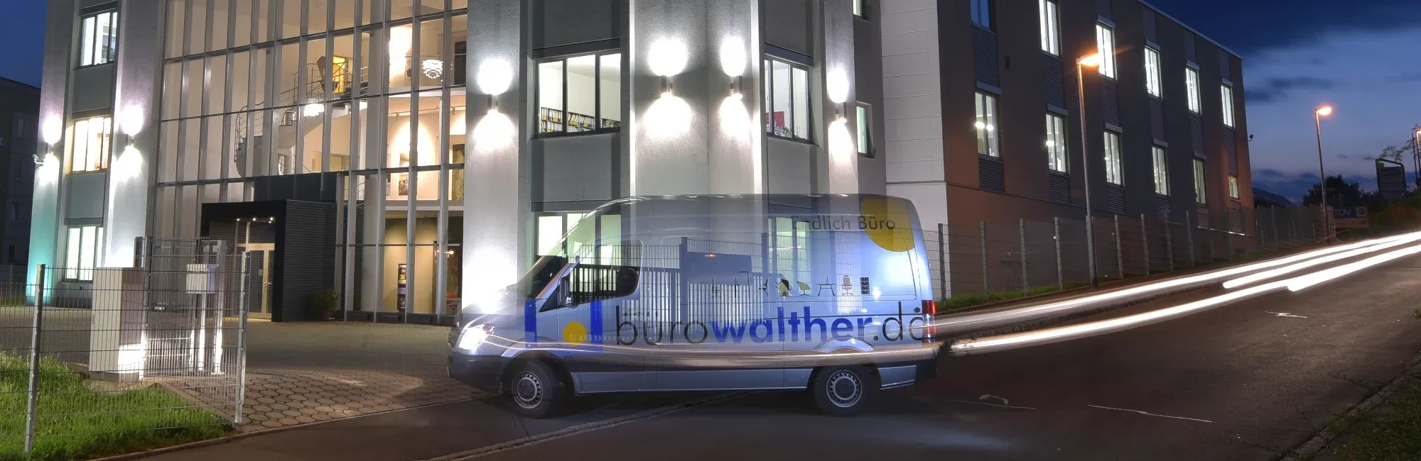 Transporter mit Aufschrift "burowalther.de" und "Endlich Büro" vor beleuchtetem Bürogebäude in der Abenddämmerung.