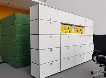 Weißer USM-Haller-Schrank mit gelben Einlegern neben gestapelten grünen Akustikelementen