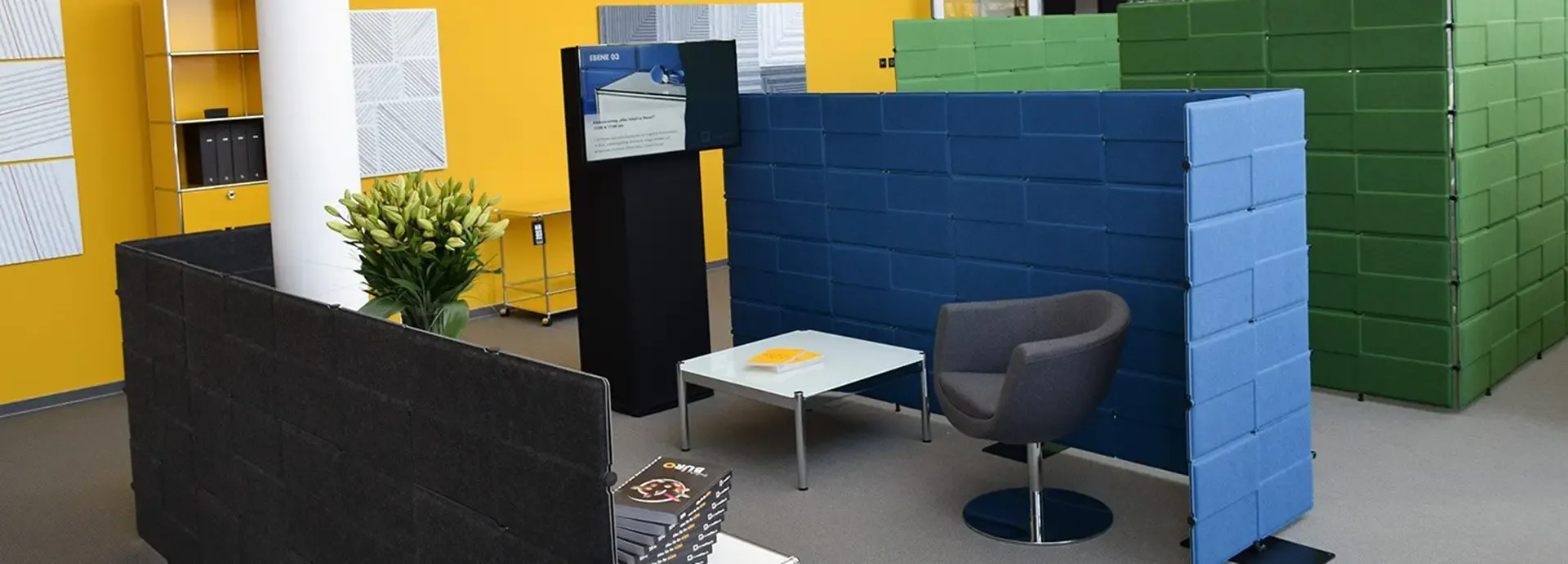 Foyer mit Akustikstellwänden, Sitzgruppe und Infostele vor gelber Wand