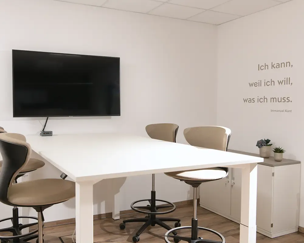 Besprechungsraum mit weißem Tisch, Bildschirm und Zitat an der Wand