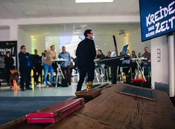 Alte Schulbank mit Tintenfass im Vordergrund, dahinter ein Redner vor dem Publikum