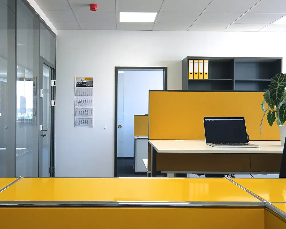 Arbeitsplatz mit gelben Trennwänden und Notebook, dahinter ein Regal