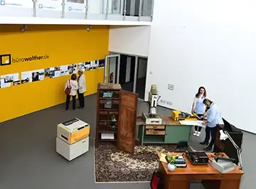 Blick von oben auf die Ausstellung historischer Bürotechnik vor der gelben Zeitwand