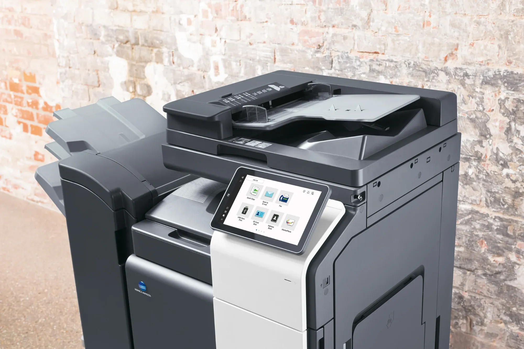 Konica Minolta Multifunktionsdrucker mit Touchscreen-Menü "Quick Copy", "Scan to E-mail", "Fax" vor Ziegelwand.