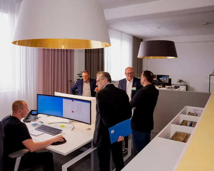 Besuchergruppe an einem Arbeitsplatz mit großem Bildschirm unter goldenen Pendelleuchten