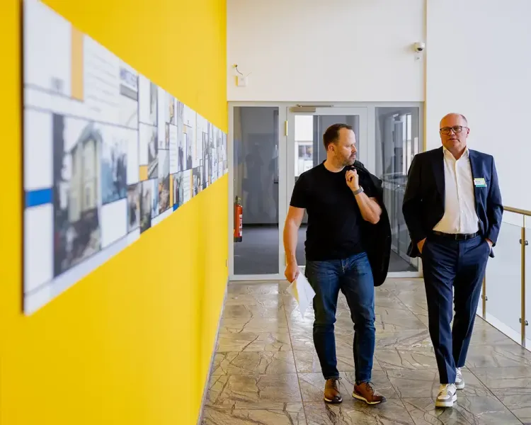 Zwei Männer gehen durch den Flur, links die gelbe Wand mit der Bilderleiste