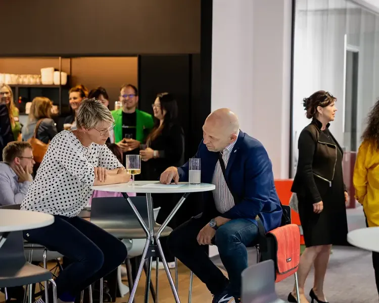 Zwei Gäste im Gespräch an einem Stehtisch, ringsum weitere Besucher
