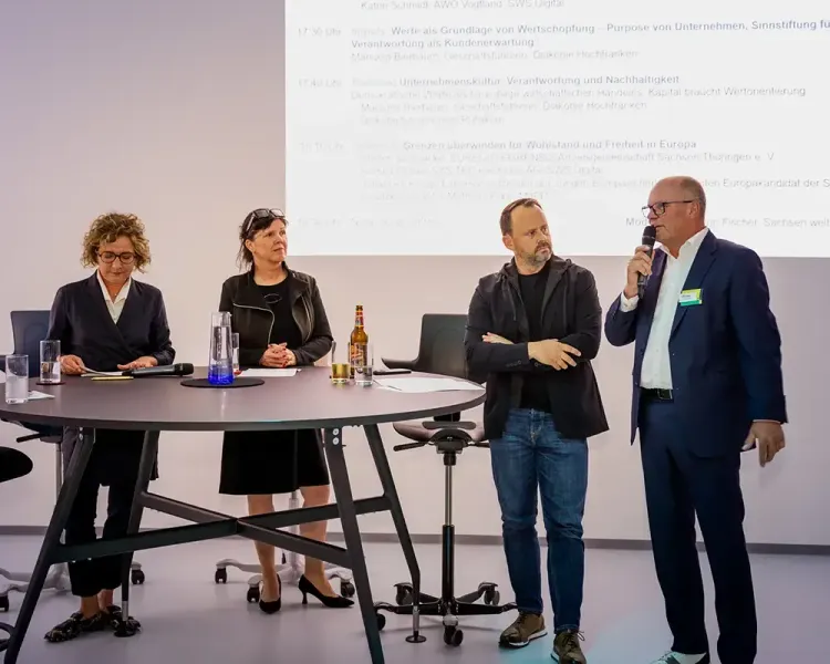 Podiumsrunde mit vier Personen vor der Leinwand, ein Redner am Mikrofon