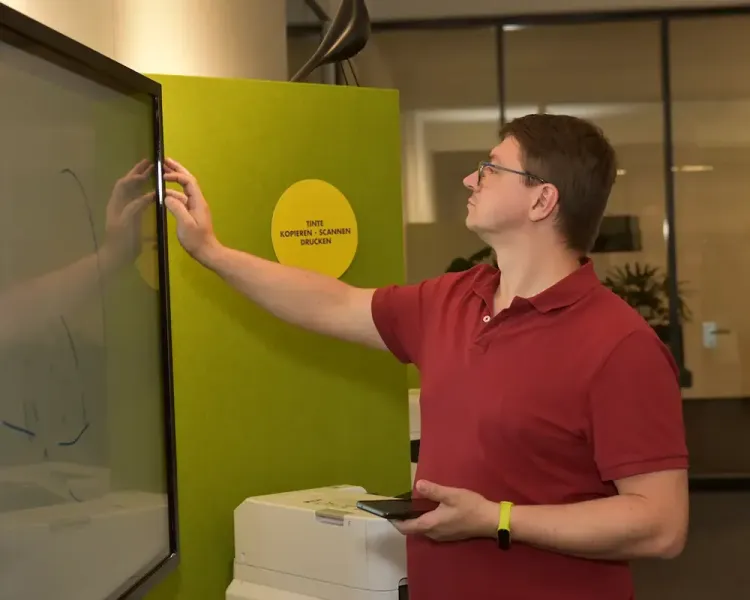 Mitarbeiter bedient ein digitales Whiteboard vor grüner Akustikwand