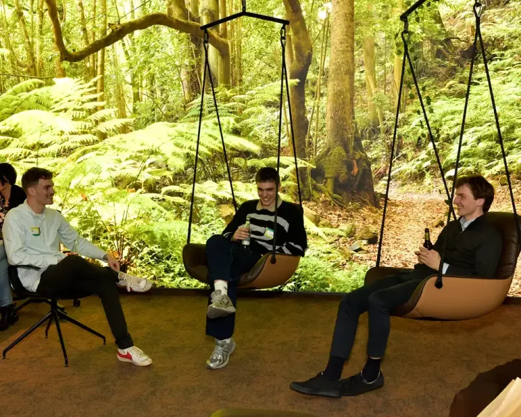 Drei junge Besucher sitzen in Hängesesseln vor der Waldtapete
