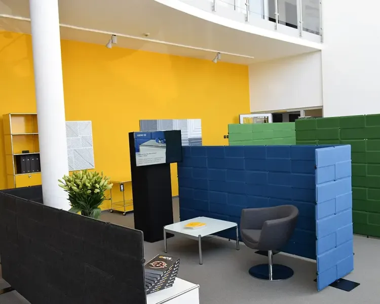 Sitzecke im Foyer mit blauen und grünen Akustikstellwänden, Sessel und Infostele vor gelber Wand