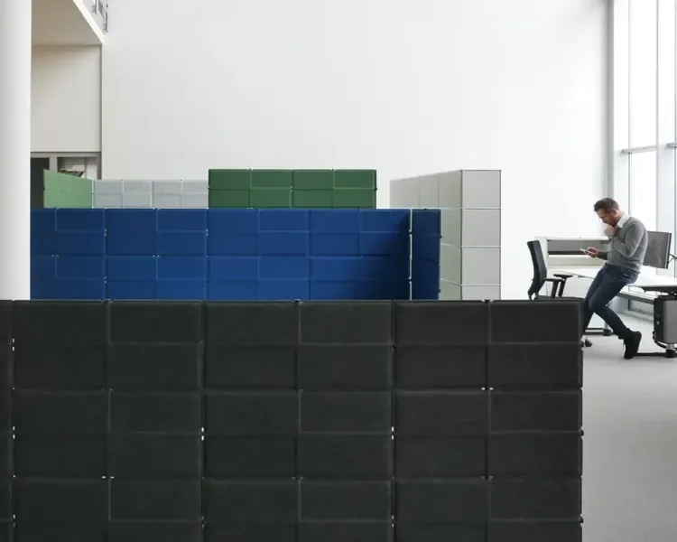 Gestapelte Akustikelemente in Schwarz, Blau, Grün und Grau vor einem Arbeitsplatz im Großraum