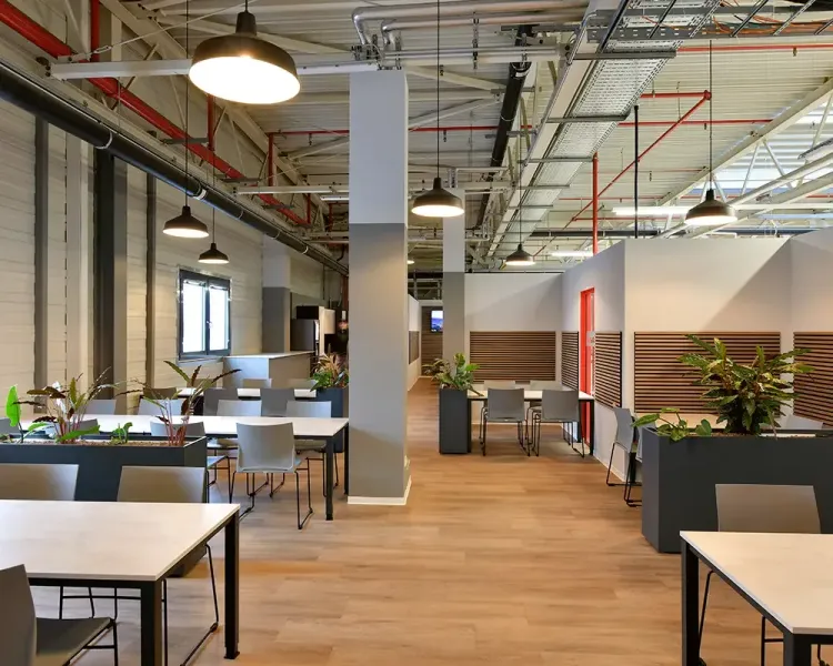 Pausenbereich in einer Halle mit Tischgruppen, Pflanzkübeln und Pendelleuchten