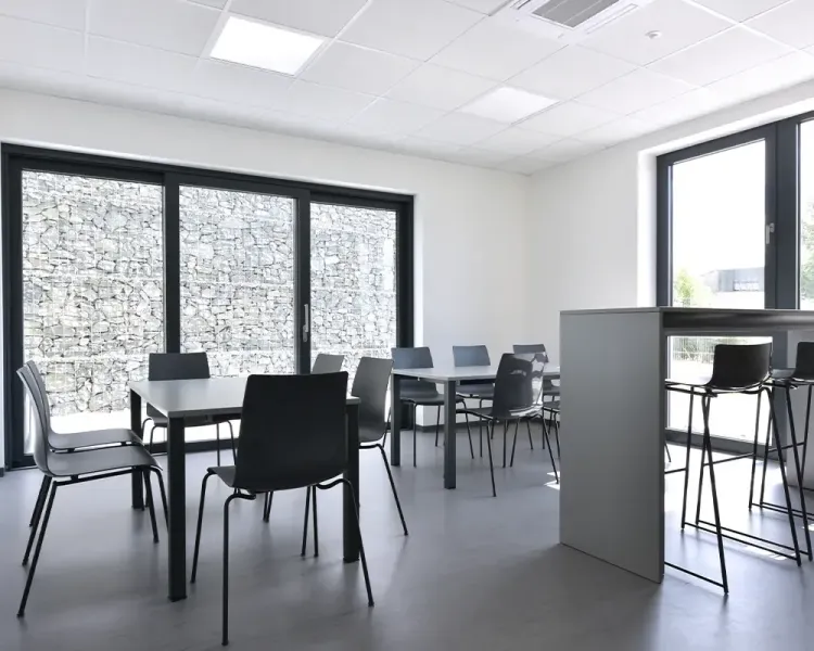 Pausenraum mit Tischgruppen und Hochtisch vor einer Gabionenwand