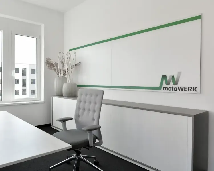 Büro mit grauem Drehstuhl vor einer Wandverkleidung mit metaWERK-Logo