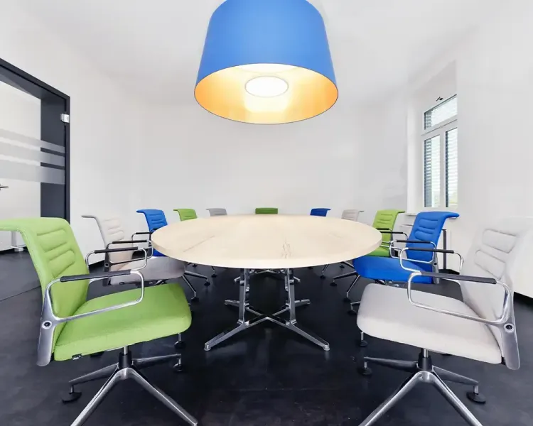 Besprechungsraum mit rundem Tisch, bunten Stühlen und blauer Pendelleuchte