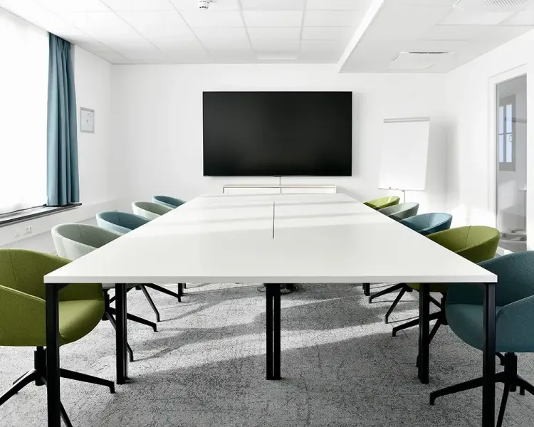 Besprechungsraum mit langer weißer Tafel, farbigen Drehsesseln und Bildschirm