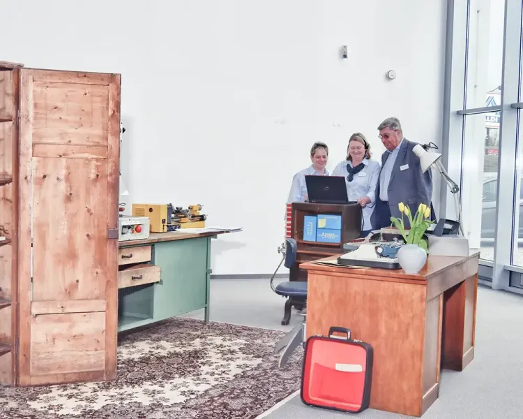 Drei Personen an einem historischen Arbeitsplatz mit Notebook und Werkbank