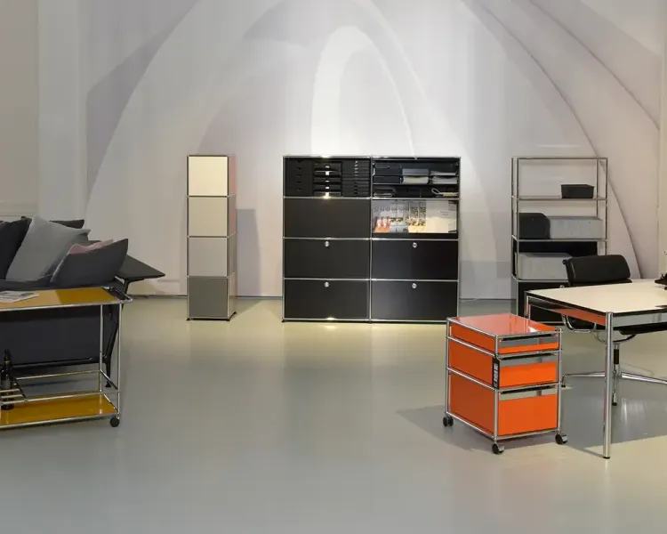 Ausstellungsraum mit schwarzem USM-Haller-Sideboard, orangefarbenem Rollcontainer und Sofa