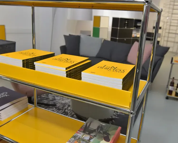 Gelbes USM-Haller-Regal mit gestapelten Büchern, dahinter ein graues Sofa