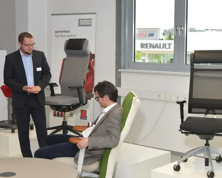 Berater erklärt einem sitzenden Besucher einen Bürodrehstuhl, daneben ein Sedus-Rollup