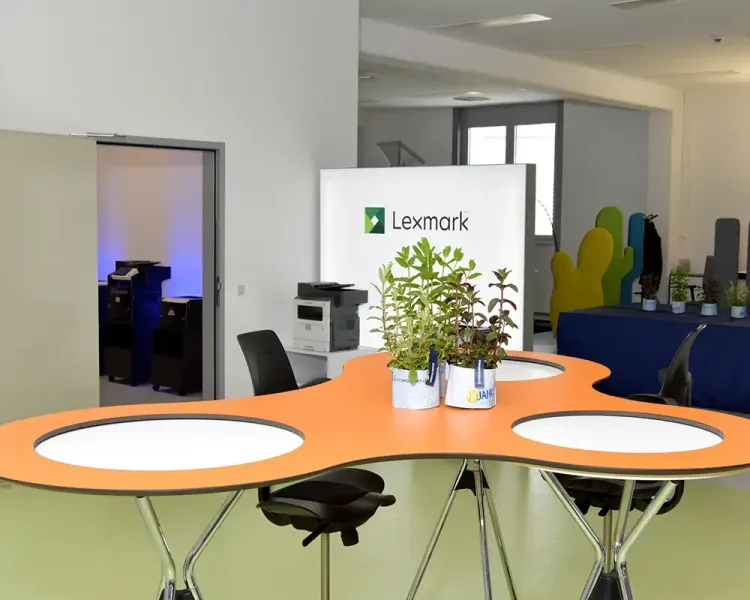 Orangefarbener Gruppentisch mit Pflanzen, dahinter ein Lexmark-Aufsteller und Drucker