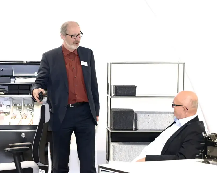 Berater erläutert einem sitzenden Gast einen Bürodrehstuhl