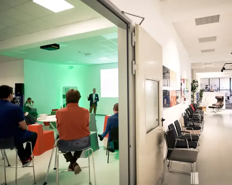 Blick vom Flur in den grün beleuchteten Vortragsraum mit Publikum