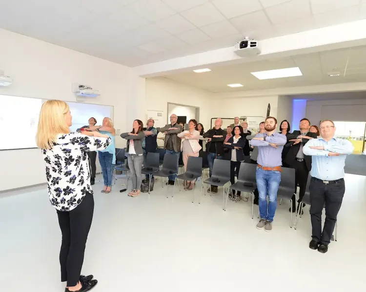 Gruppe im Schulungsraum macht unter Anleitung eine Dehnübung im Stehen