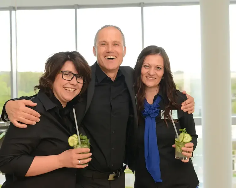 Drei Mitarbeitende mit Cocktails vor der Fensterfront