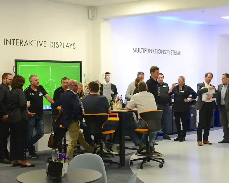 Gäste an Stehtischen zwischen den Bereichen „Interaktive Displays“ und „Multifunktionssysteme“