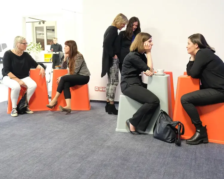 Besucherinnen sitzen auf orangefarbenen und grauen Hockern im Gespräch