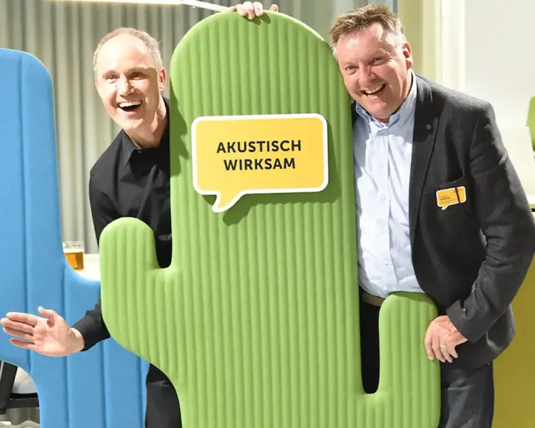 Zwei Gäste posieren hinter einem grünen Akustikkaktus mit dem Schild „Akustisch wirksam“