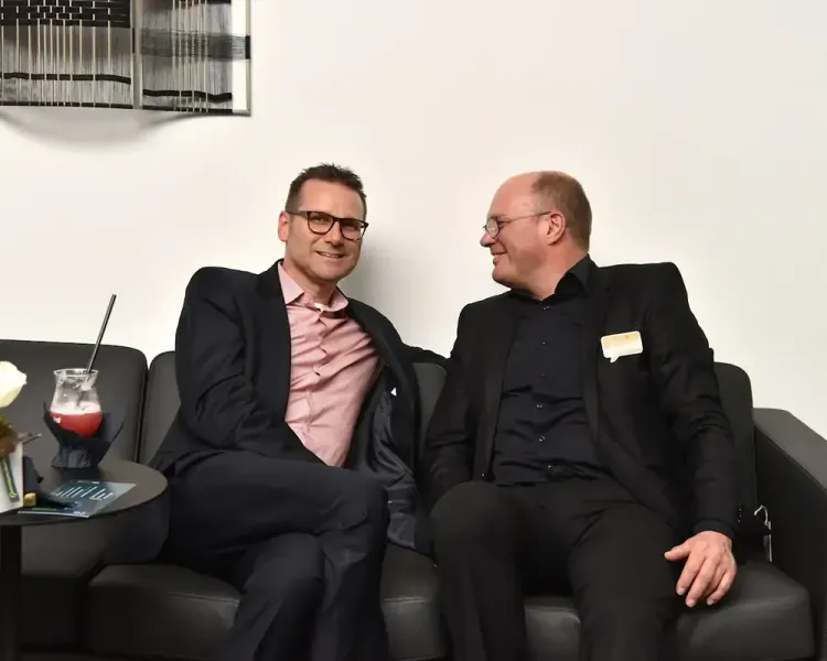 Zwei Gäste sitzen lachend auf einem schwarzen Ledersofa