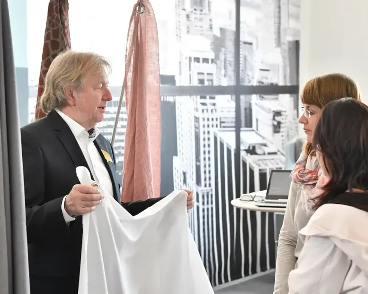 Berater zeigt zwei Besucherinnen ein weißes Textil, dahinter eine Stadtfototapete