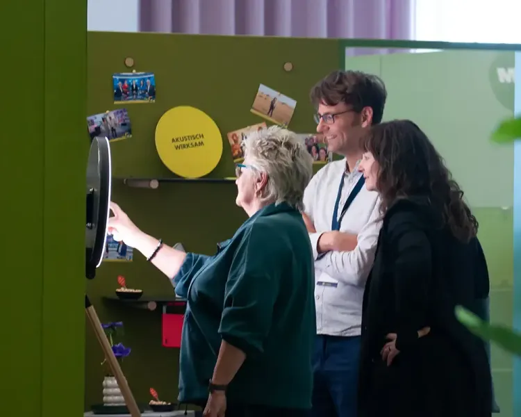 Besucherin fotografiert sich mit zwei Gästen an einer Fotobox vor grüner Wand