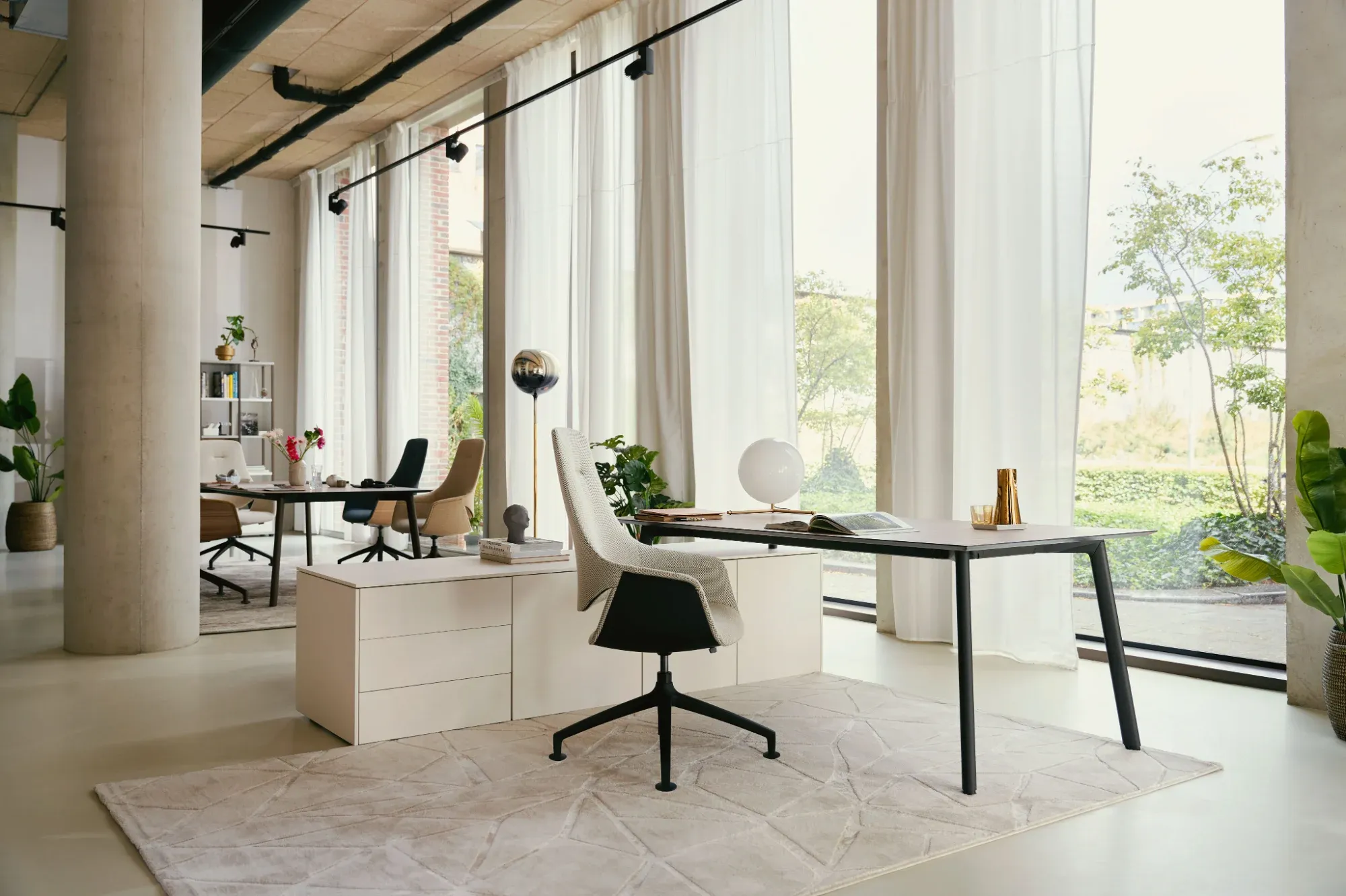 Helles Büro mit Schreibtisch, Drehstuhl, weißem Sideboard, Teppich und raumhohen Fenstern mit Vorhängen.
